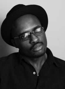 headshot of a black man wearing a dark shirt and a black hat looking at the camera