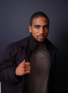 picture of a black male model wearing a dark shirt and black coat against a dark background