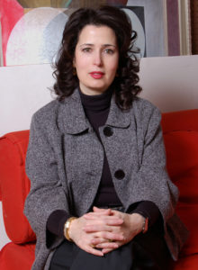 headshot of an interior designer in dark grey jacket and black sweater sitting on a red couch